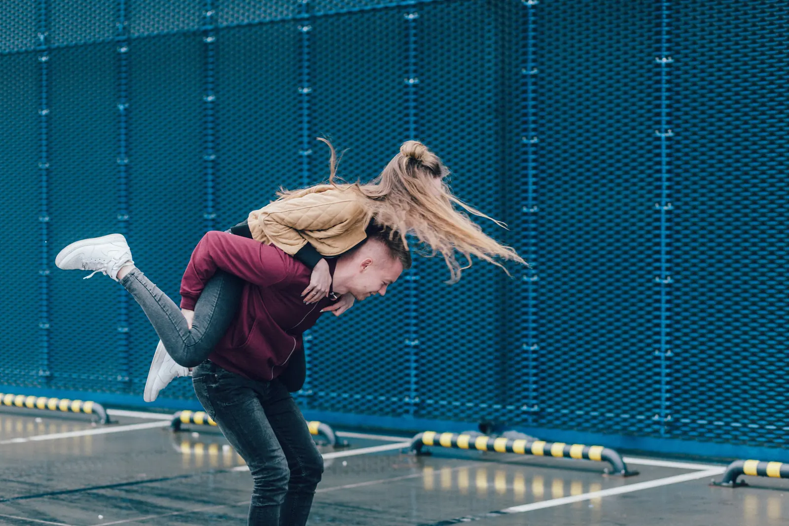 A laughing woman leaps onto her partner's back for a piggyback hug in front of a blue mesh wall