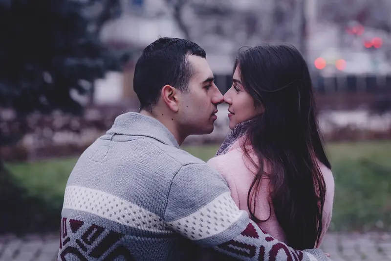 A couple in cozy sweaters leans in close, about to kiss, outdoors on a gray day