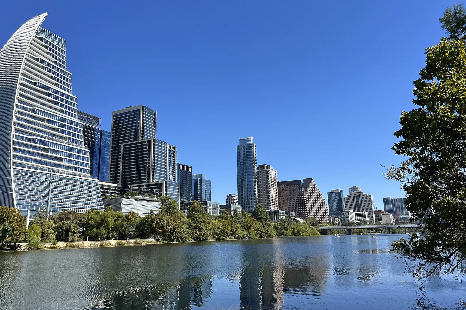 Downtown Austin's high-rises, including a curved glass tower, reflect in Lady Bird Lake under a clear blue sky