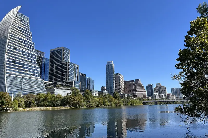 Downtown Austin's high-rises, including a curved glass tower, reflect in Lady Bird Lake under a clear blue sky