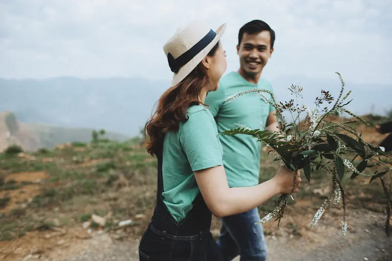 A smiling couple in matching teal shirts walks across a grassy hillside, she in a sun hat holding a bundle of greenery