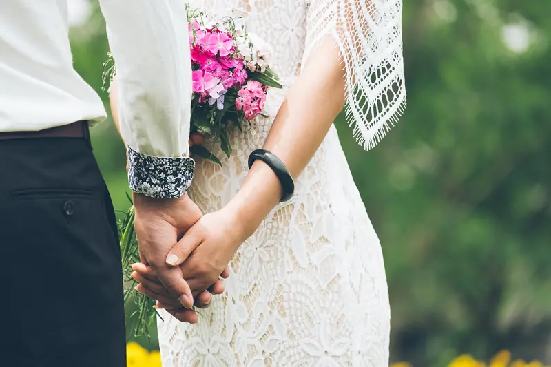 A bride in a lace dress holding a pink and white bouquet clasps hands with her groom