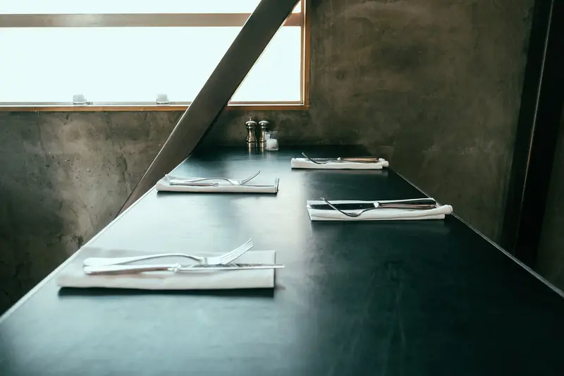 Folded white cloth napkins with forks and knives are set along a long dark table by a window