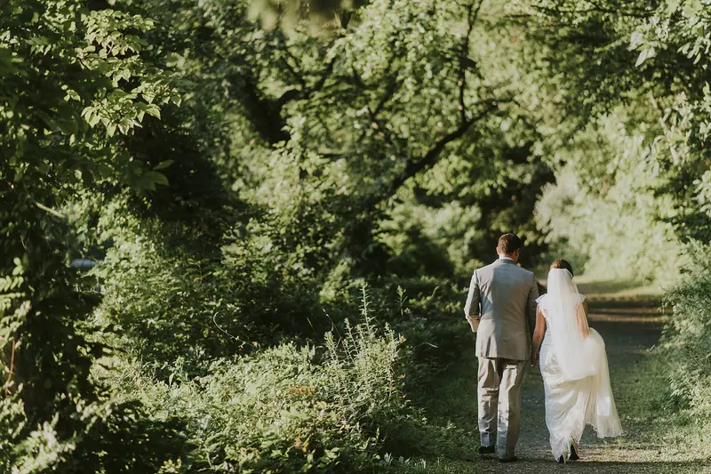 A bride and groom walk away together down a sunlit path through lush green woods