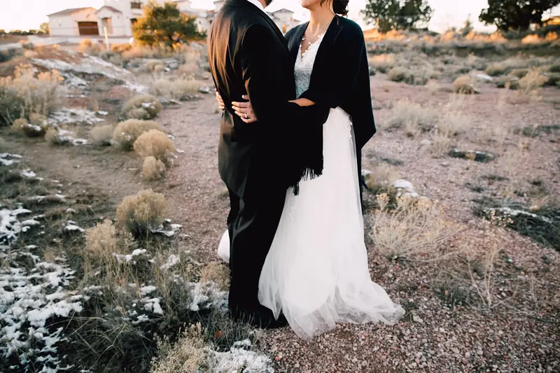 A bride in a white gown with a dark shawl and her groom embrace on a scrubby desert trail
