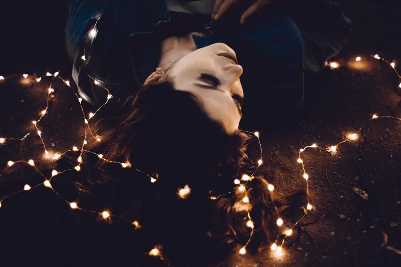 A young woman lies on the ground at night surrounded by glowing strands of warm fairy lights
