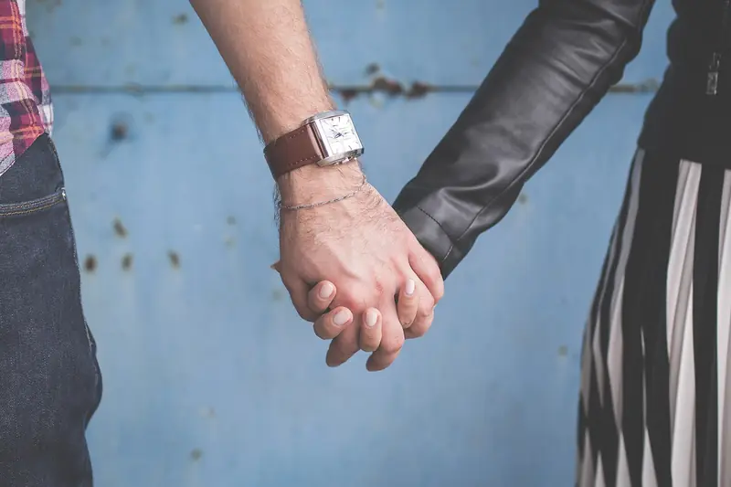 A couple in casual layered outfits holds hands in front of a weathered blue wall