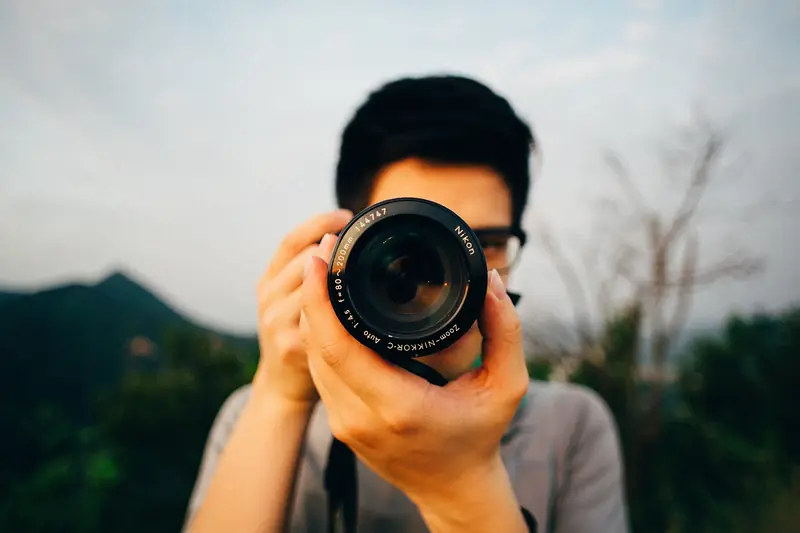 A photographer holds a DSLR up to their eye and aims it straight at the viewer, with hills blurred behind