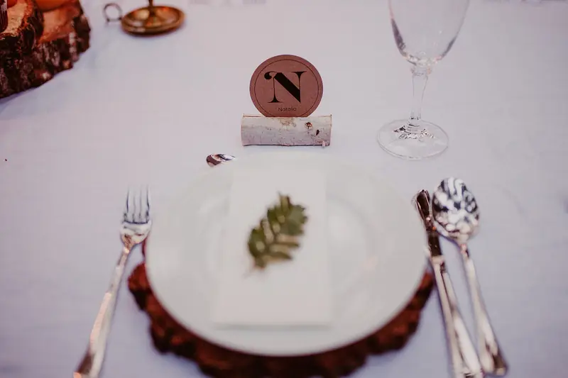 A wedding place setting with a white plate, a sprig of greenery, silverware and a round monogram card on a wood slice