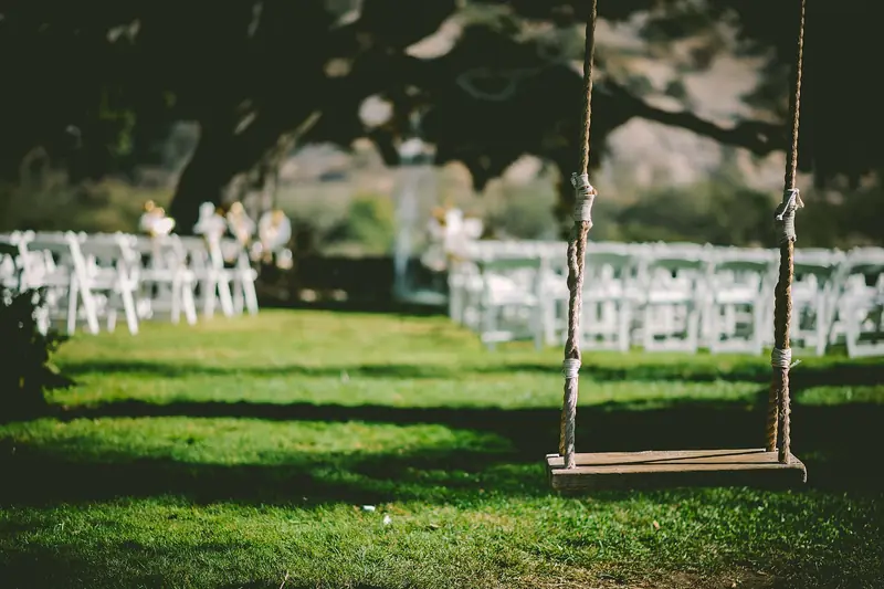 A wooden rope swing hangs from a tree over a green lawn set with rows of white wedding ceremony chairs