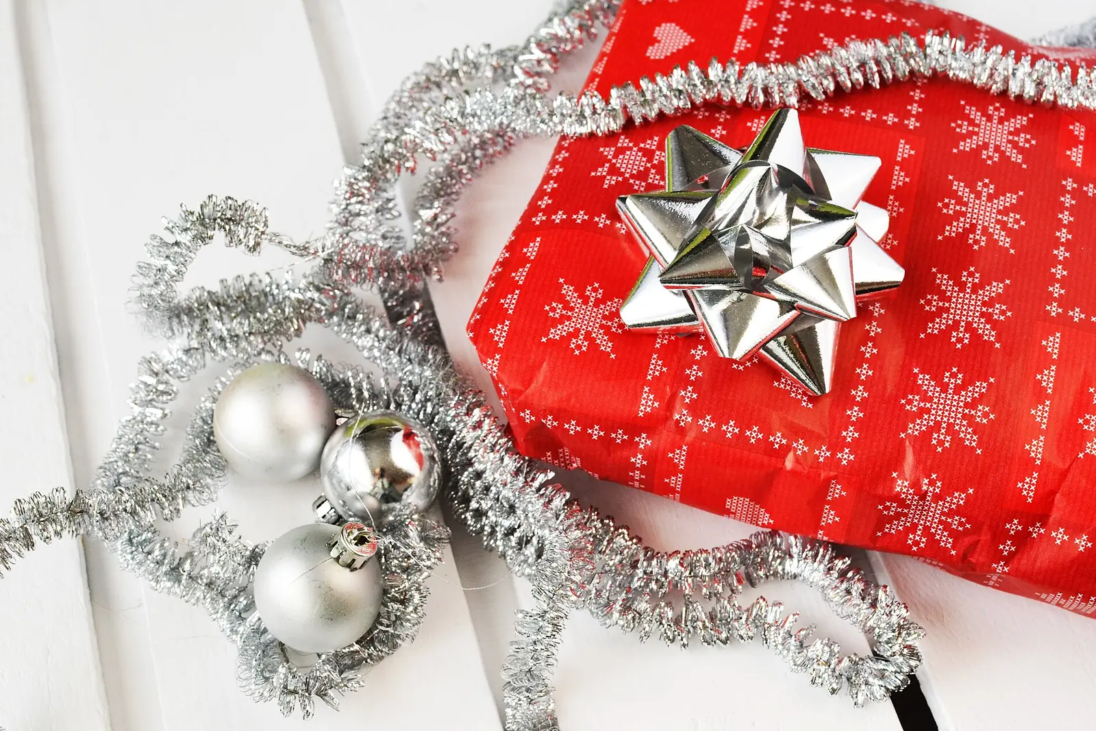 A gift wrapped in red snowflake paper with a silver bow sits beside silver ornaments and tinsel on white wood