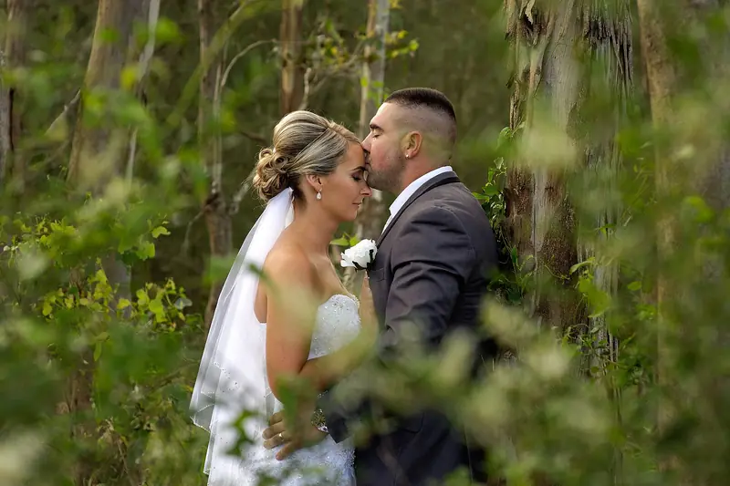 A groom kisses his bride's forehead in a lush, green wooded garden