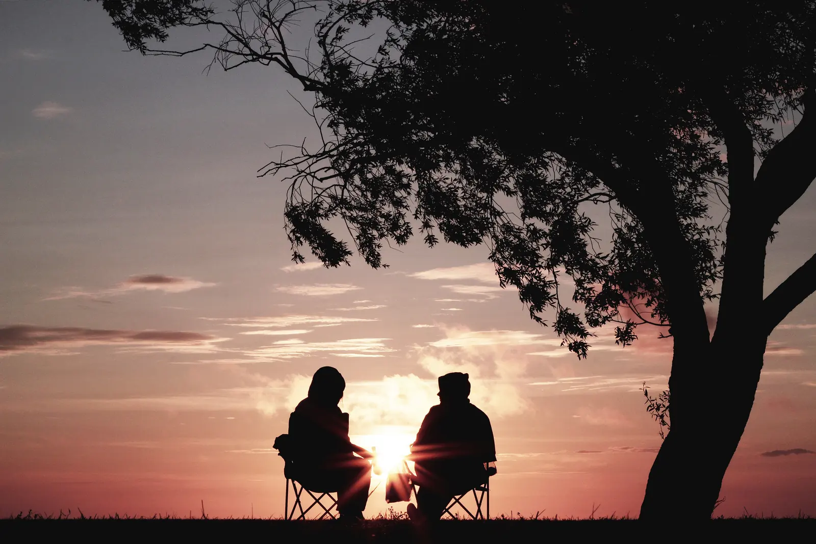 A couple sits on chairs in silhouette beneath a tree, watching a glowing pink and orange sunset