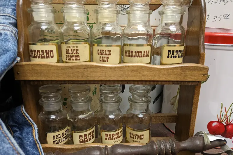 A vintage wooden spice rack holds glass jars with gold labels reading Oregano, Garlic Salt, Marjoram, Sage, Parsley and Thyme
