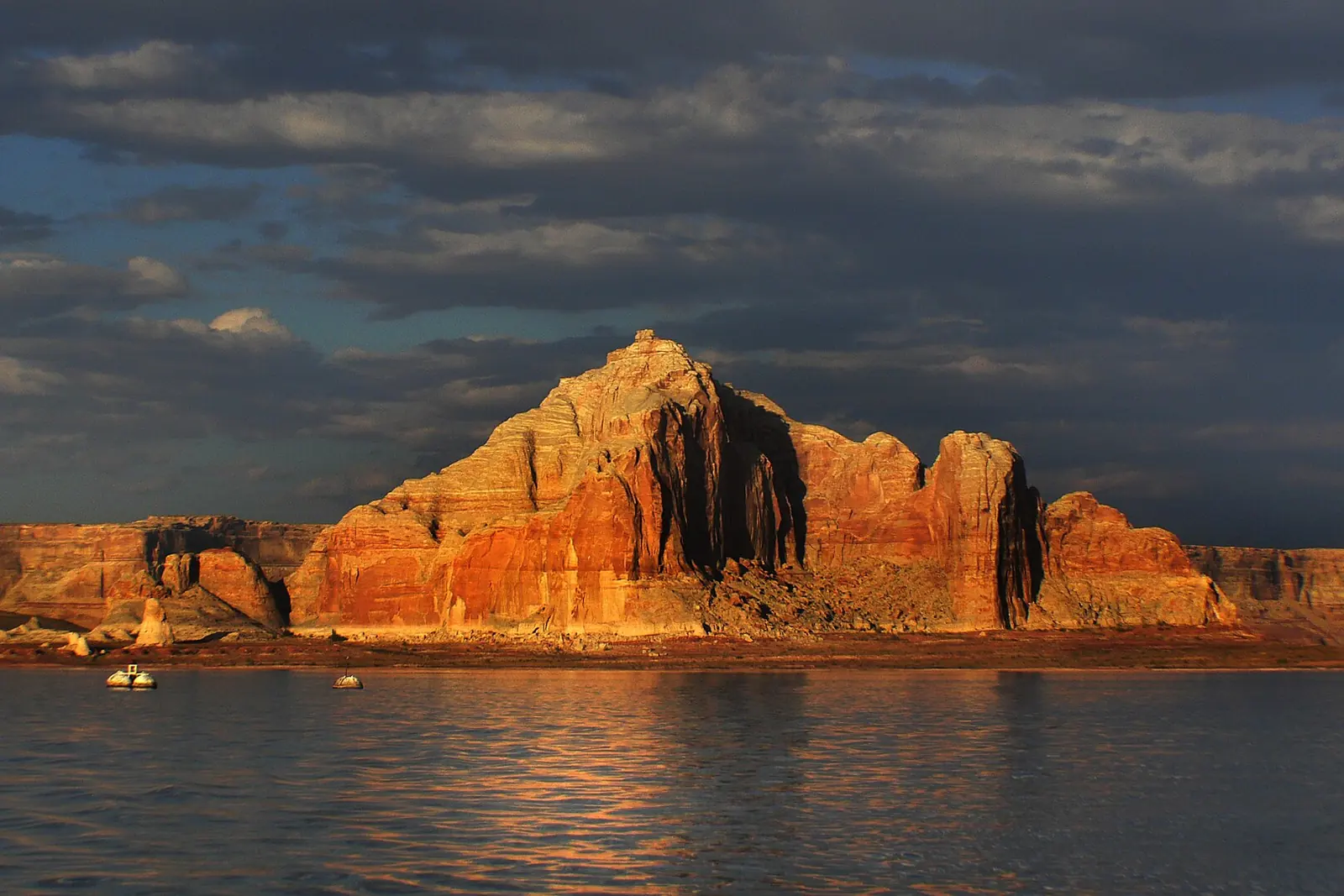 A red sandstone butte lit by golden light rises above the calm water of Lake Powell under a dark sky