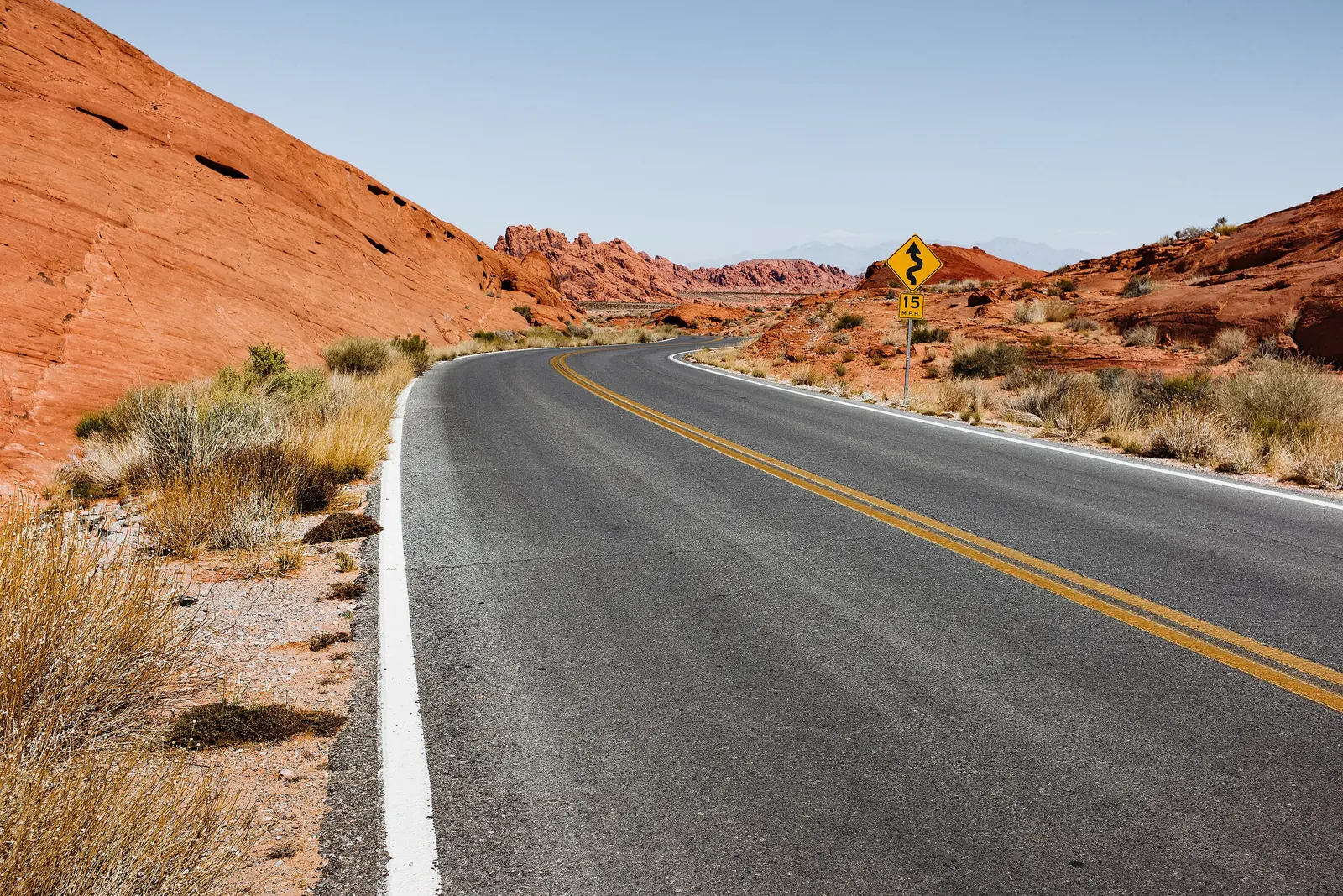 A two-lane road curves through red rock desert hills under a clear blue sky