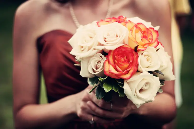 A bridesmaid in a rust-colored dress holds a bouquet of cream and orange roses