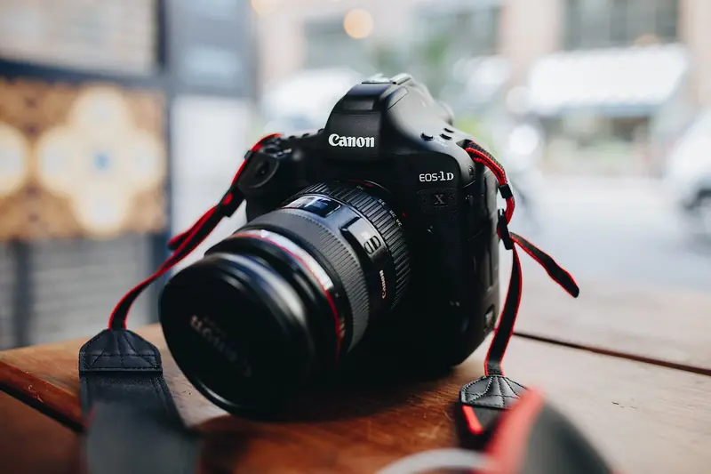 A Canon DSLR with a large zoom lens and red strap sits on a wooden table