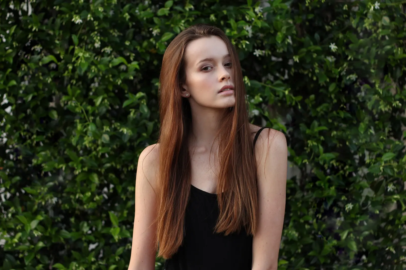 A young woman with long auburn hair and a black top poses in front of a wall of green leaves