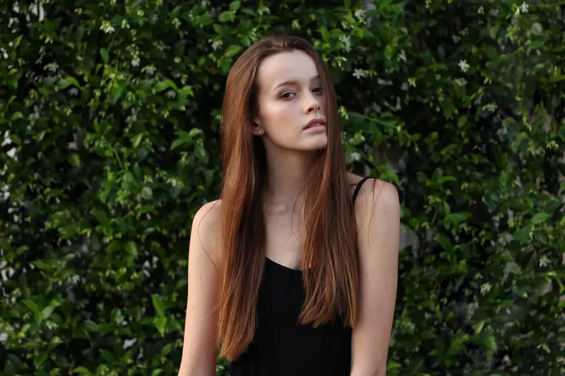 A young woman with long auburn hair and a black top poses in front of a wall of green leaves