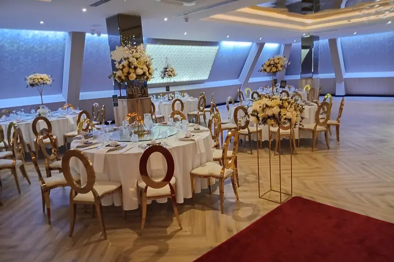 A banquet hall set for a reception with round white-draped tables, gold chairs and tall floral centerpieces