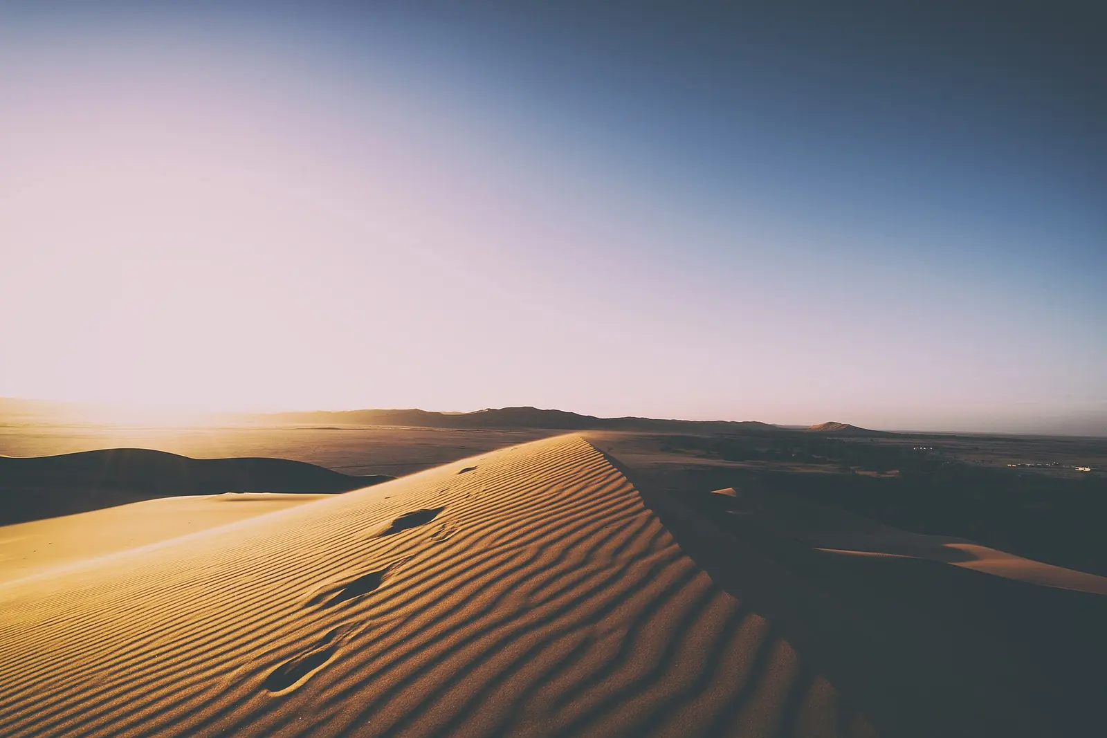 Rippled sand dunes glow in warm evening light under a pastel sky, with footprints along the crest