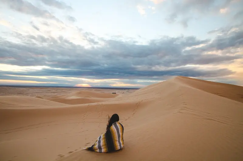 A person wrapped in a striped blanket sits alone among rolling sand dunes under a wide cloudy sky