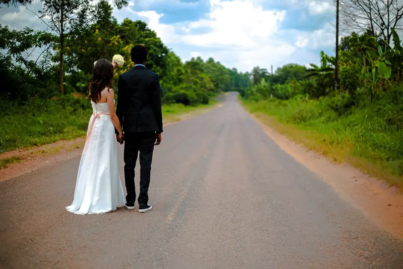 A bride and groom stand hand in hand on a quiet rural road lined with green trees