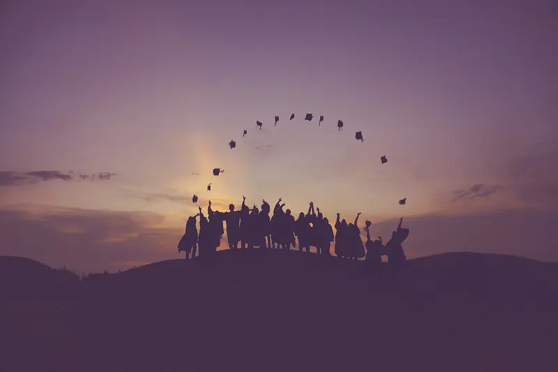 Silhouetted graduates toss their caps into a purple sunset sky on a hilltop