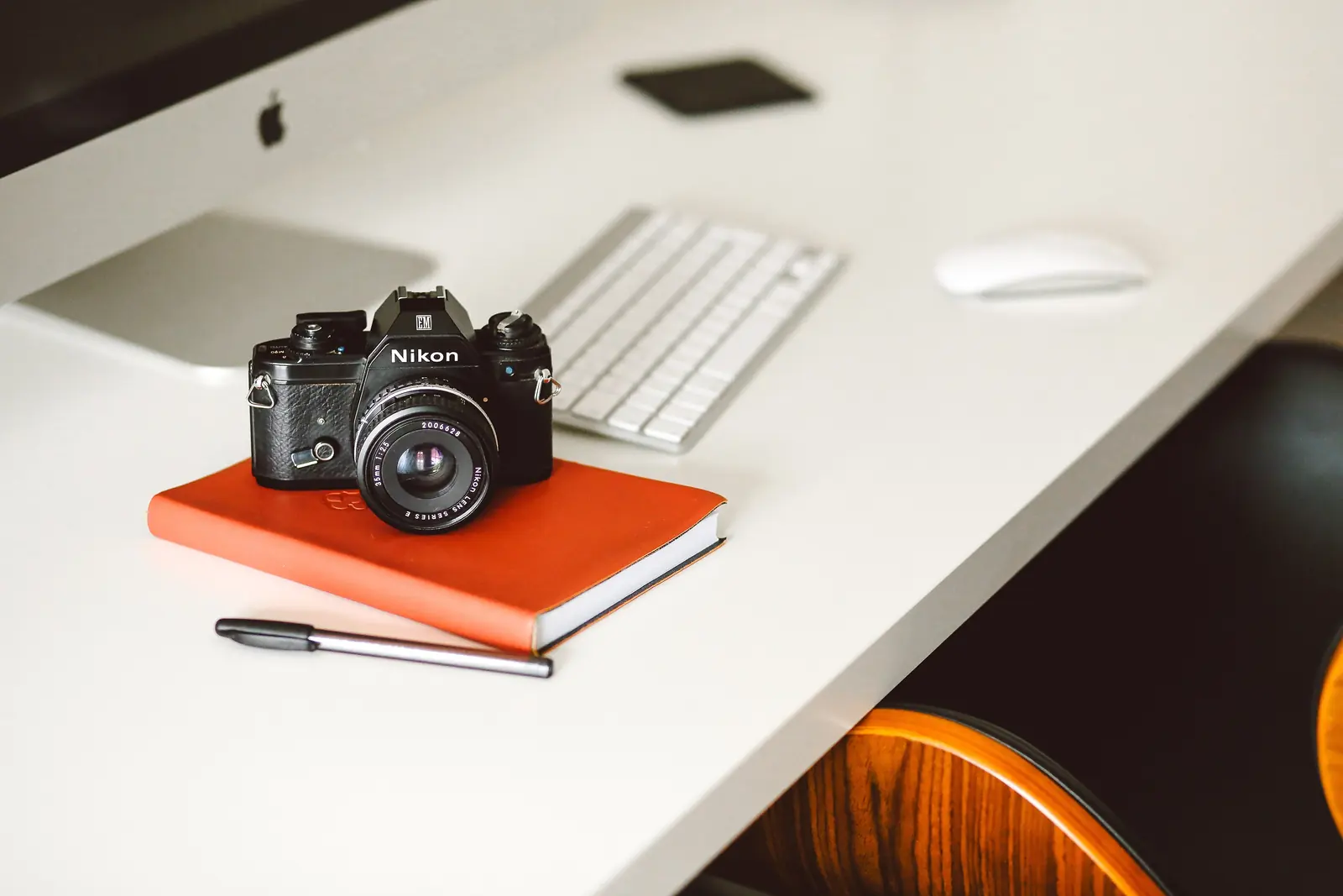 A black Nikon film camera rests on an orange notebook on a white desk beside a laptop keyboard