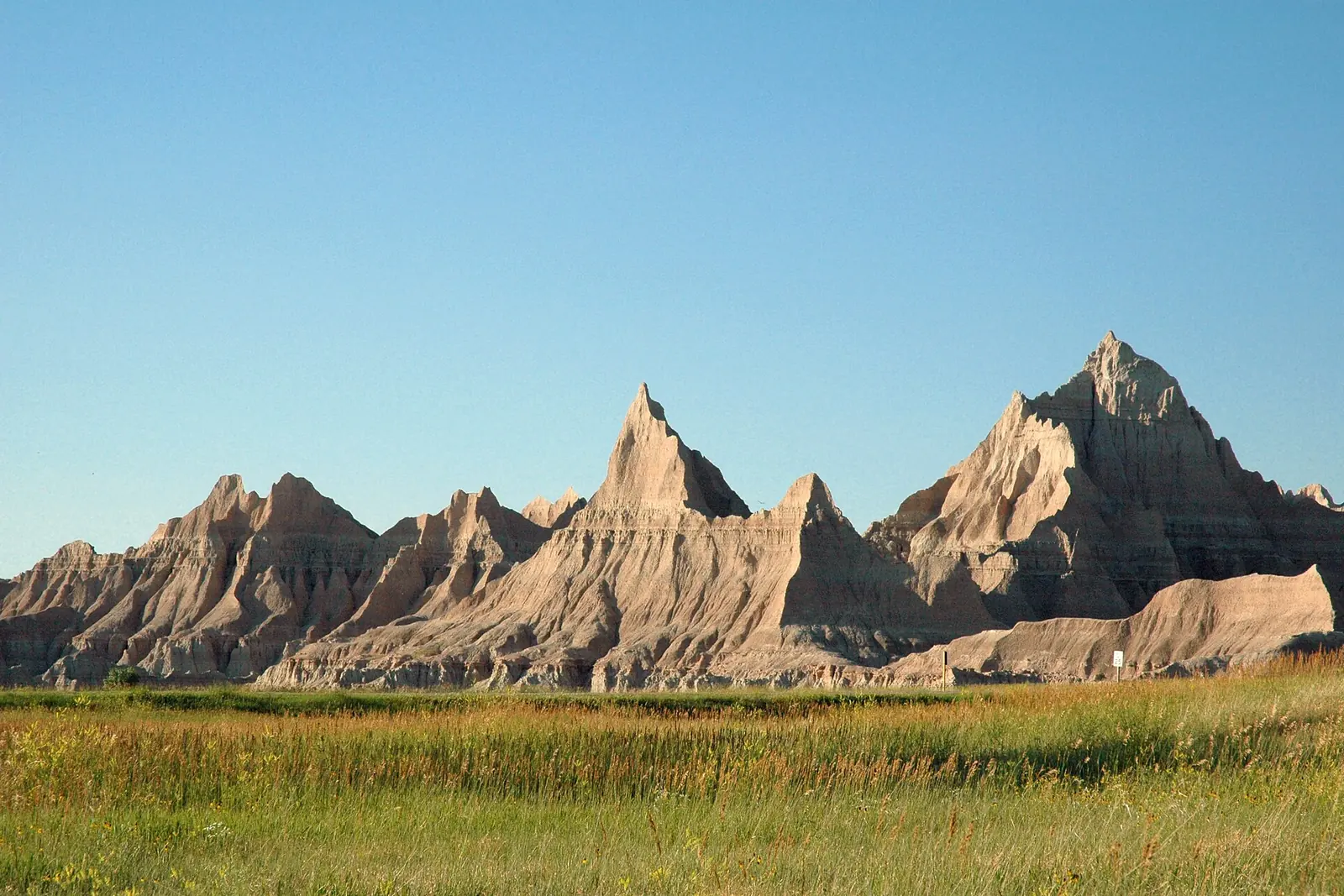 Jagged, eroded spires of Badlands National Park rise above a green prairie under a clear blue sky