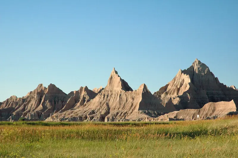 Jagged, eroded spires of Badlands National Park rise above a green prairie under a clear blue sky