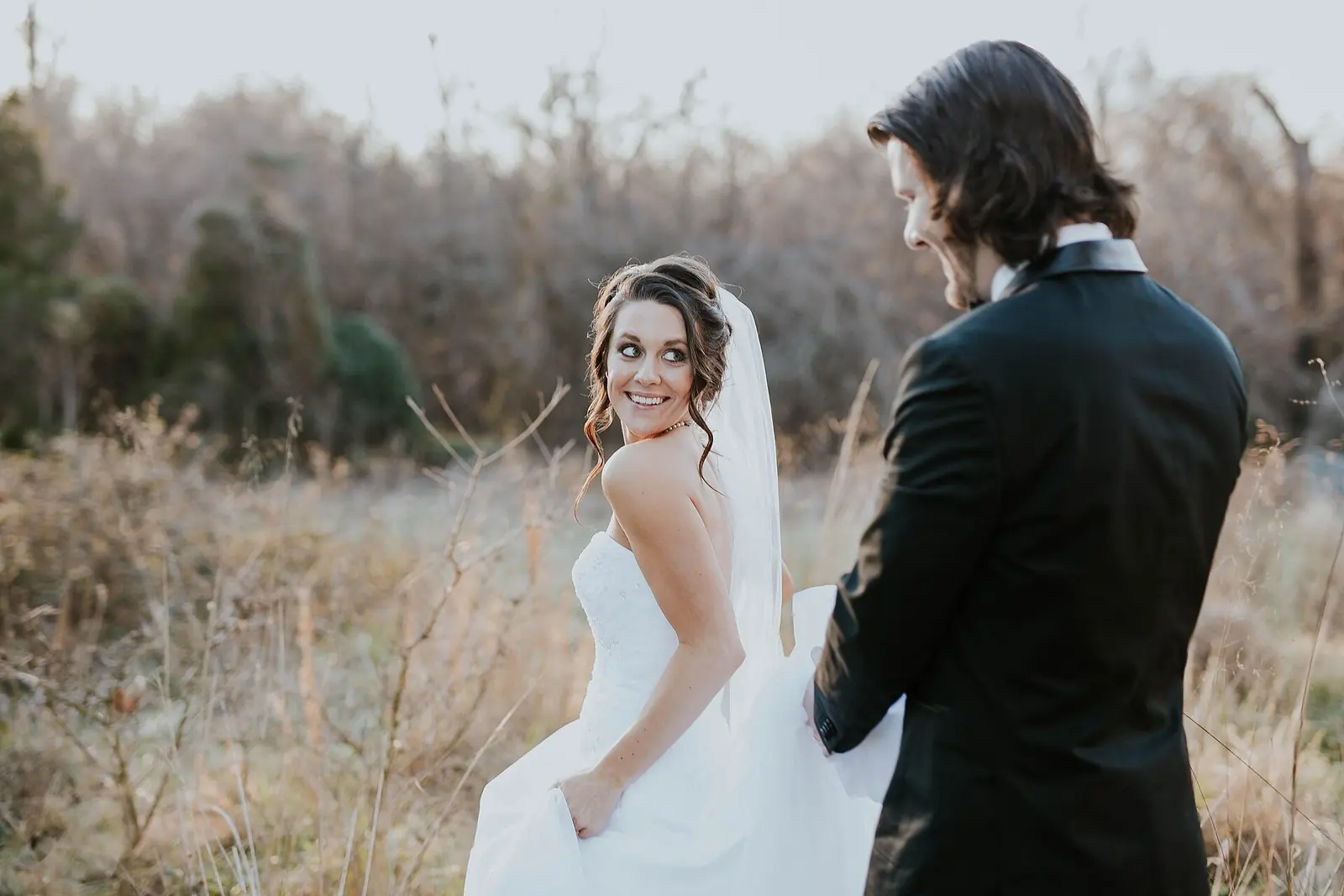 A smiling bride glances back over her shoulder at her groom in a golden autumn field