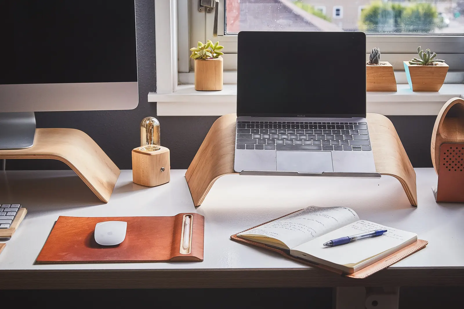 An open notepad and pen sit on a tidy desk with a laptop, monitor and small potted plants