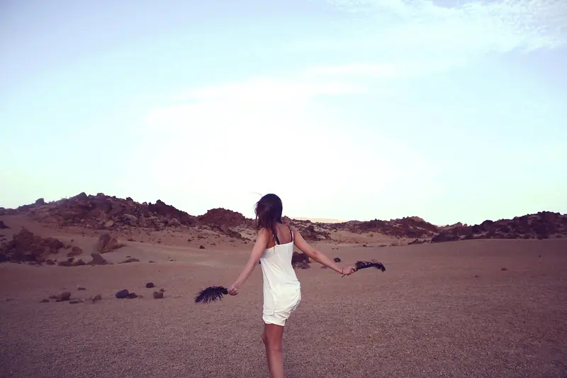 A young woman in a white dress dances with arms outstretched on a rocky desert plain at dusk