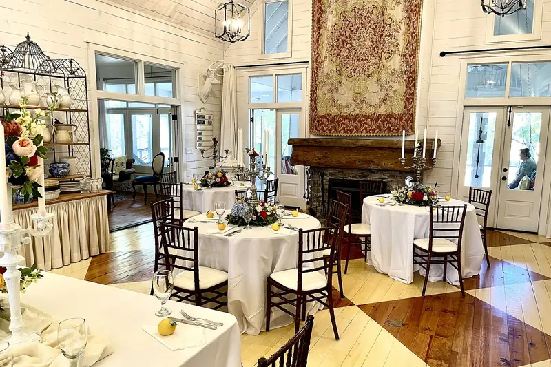 A bright wedding venue interior with white-clothed reception tables, chandeliers and a fireplace
