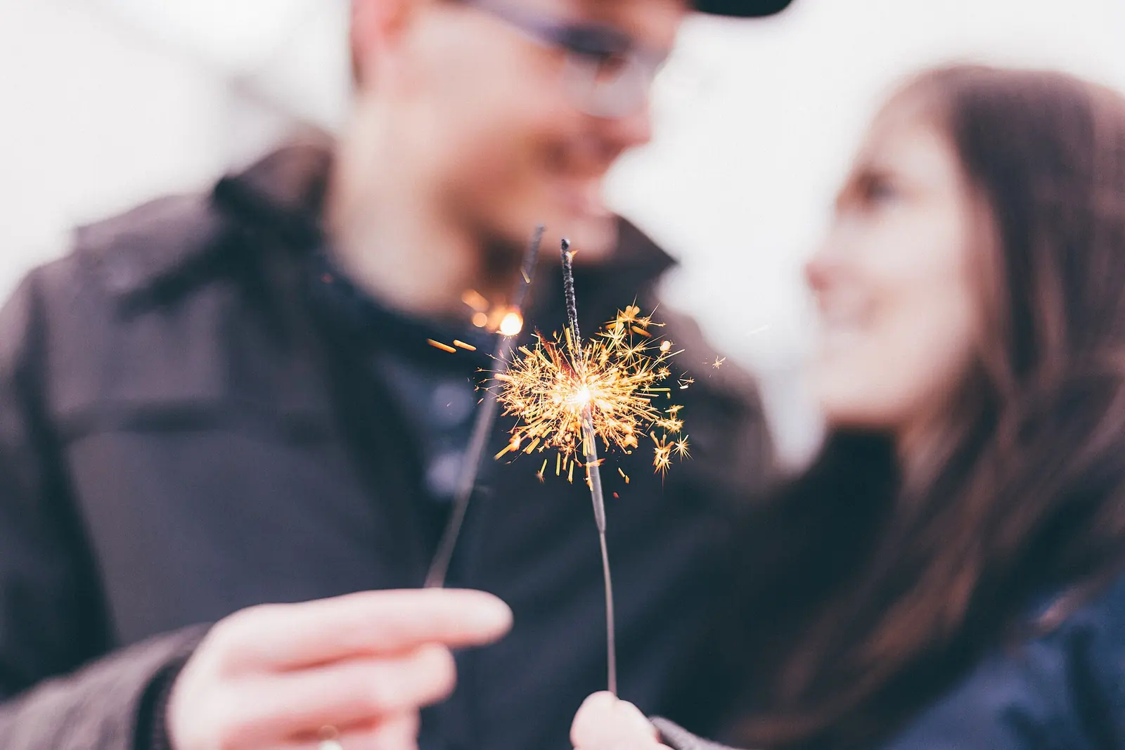 A smiling couple holds lit sparklers close together, the sparks glowing between them