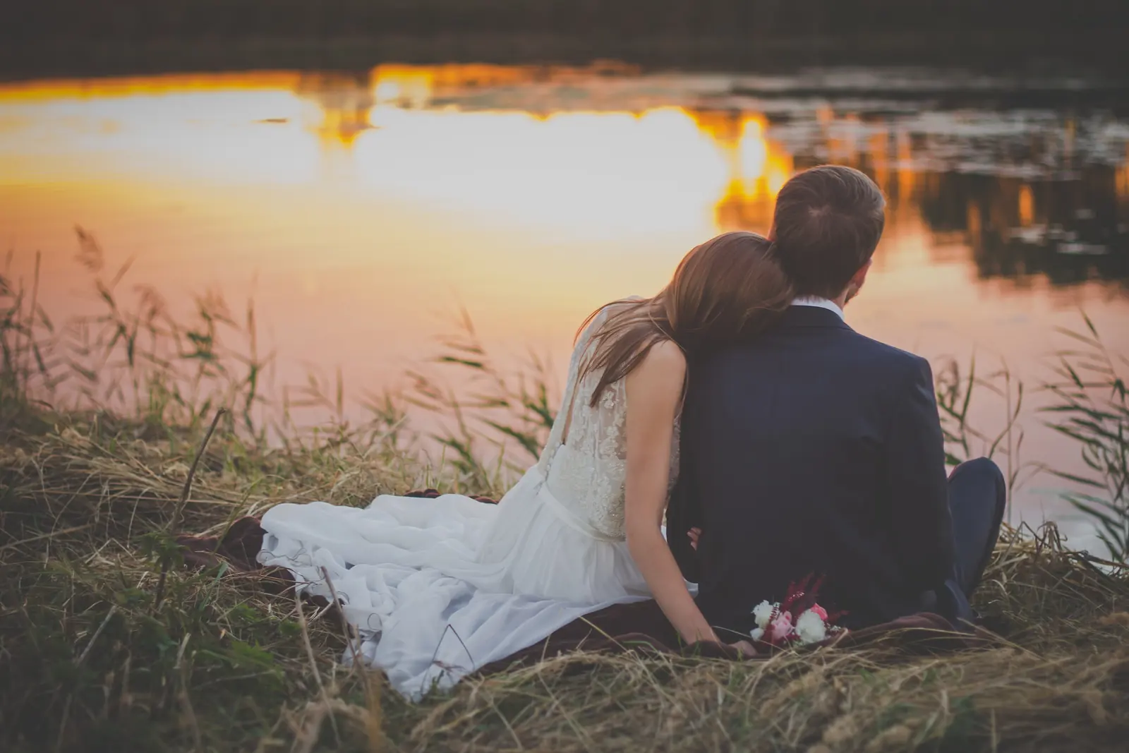 A bride and groom sit close together in tall grass by a pond, watching a golden sunset reflected in the water
